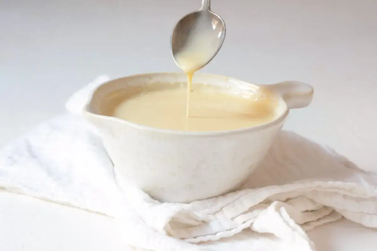 Crema inglesa cayendo de una cuchara a un cuenco blanco, sobre una tela arrugada.