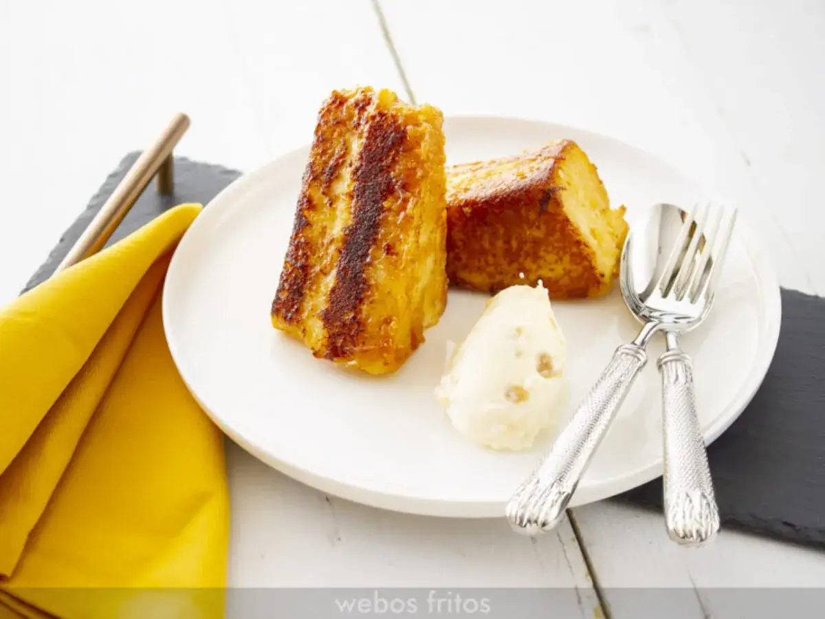 Dos porciones doradas de pan brioche para torrijas, acompañadas de una bola de helado y cubiertos plateados.