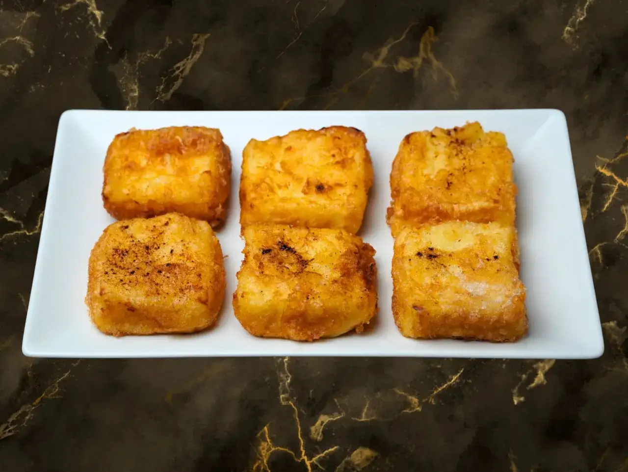 Seis porciones doradas de leche frita, preparadas con Thermomix, servidas en plato blanco sobre fondo oscuro.
