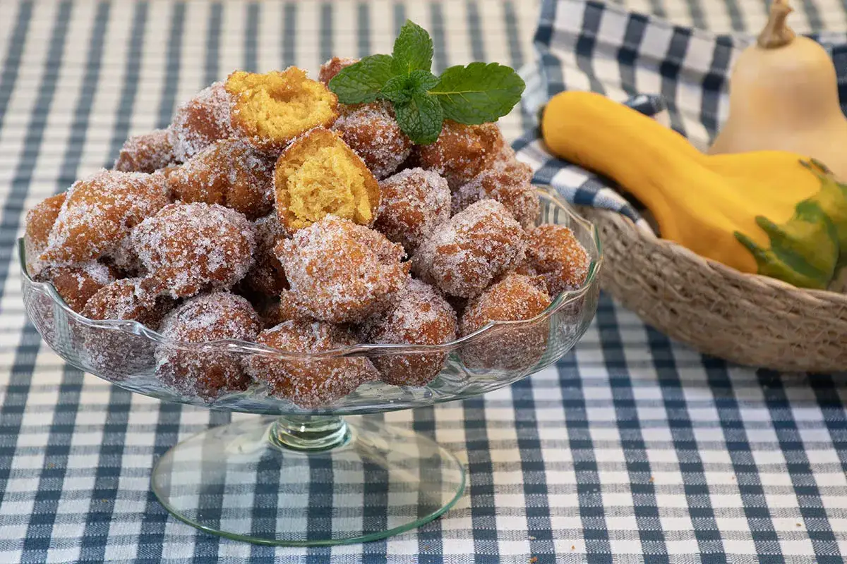 Un tazón lleno de buñuelos de calabaza cubiertos de azúcar, adornados con una ramita de menta. Al fondo, calabazas en una cesta.