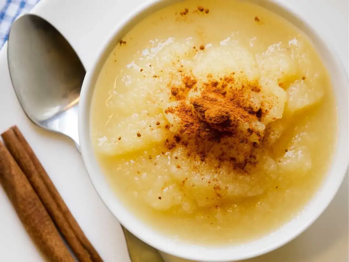 Un tazón de compota de manzana casera, espolvoreada con canela y acompañada de dos ramas de canela y una cuchara.
