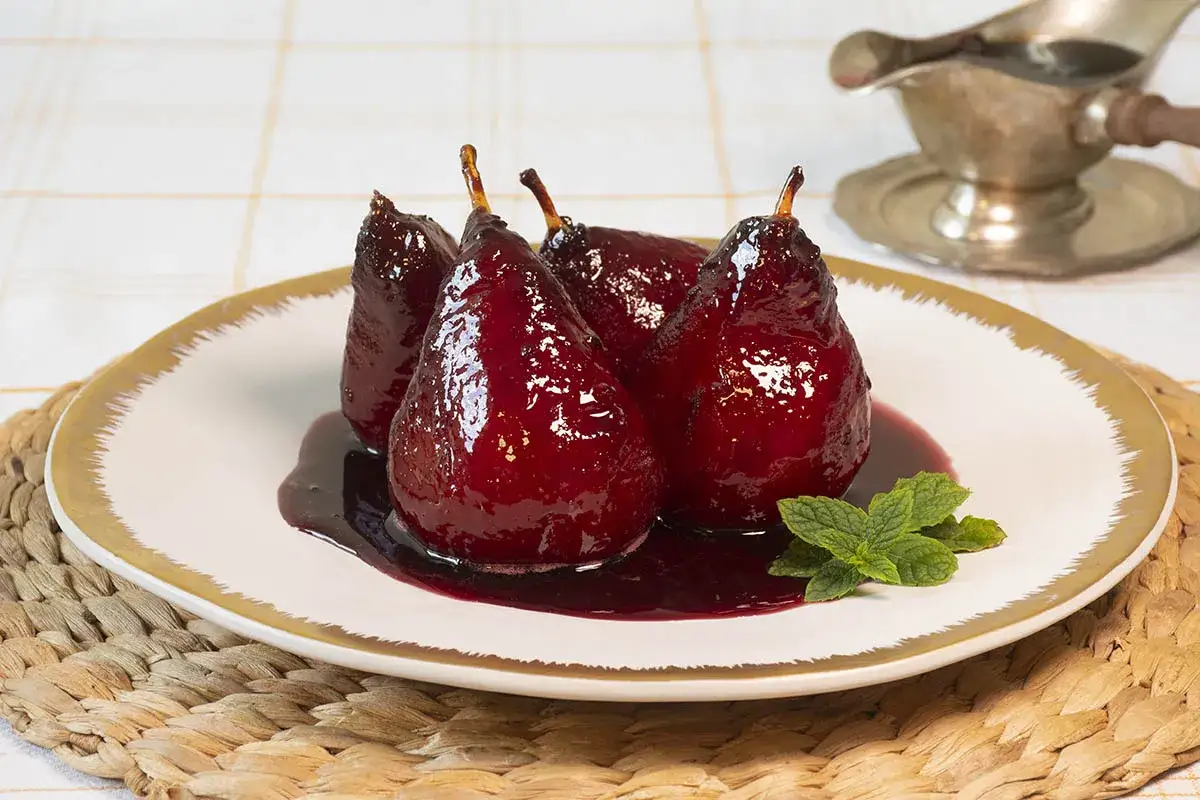 Cuatro peras al vino, brillantes y oscuras, reposan en un plato blanco con borde dorado, bañadas en su propio almíbar rojizo. Una ramita de menta fresca añade un toque de color.