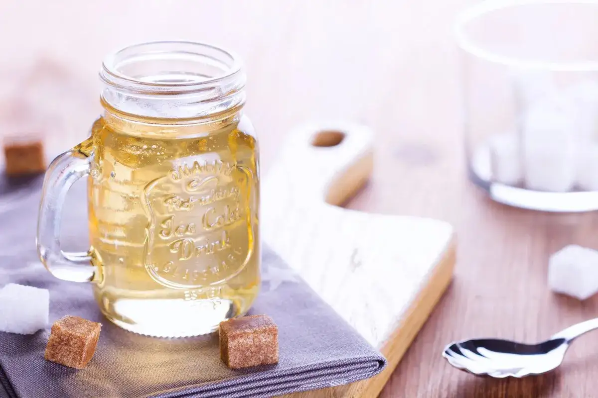 Un tarro de cristal con líquido dorado, rodeado de terrones de azúcar. Podría ser un delicioso jarabe de azúcar invertida para repostería.