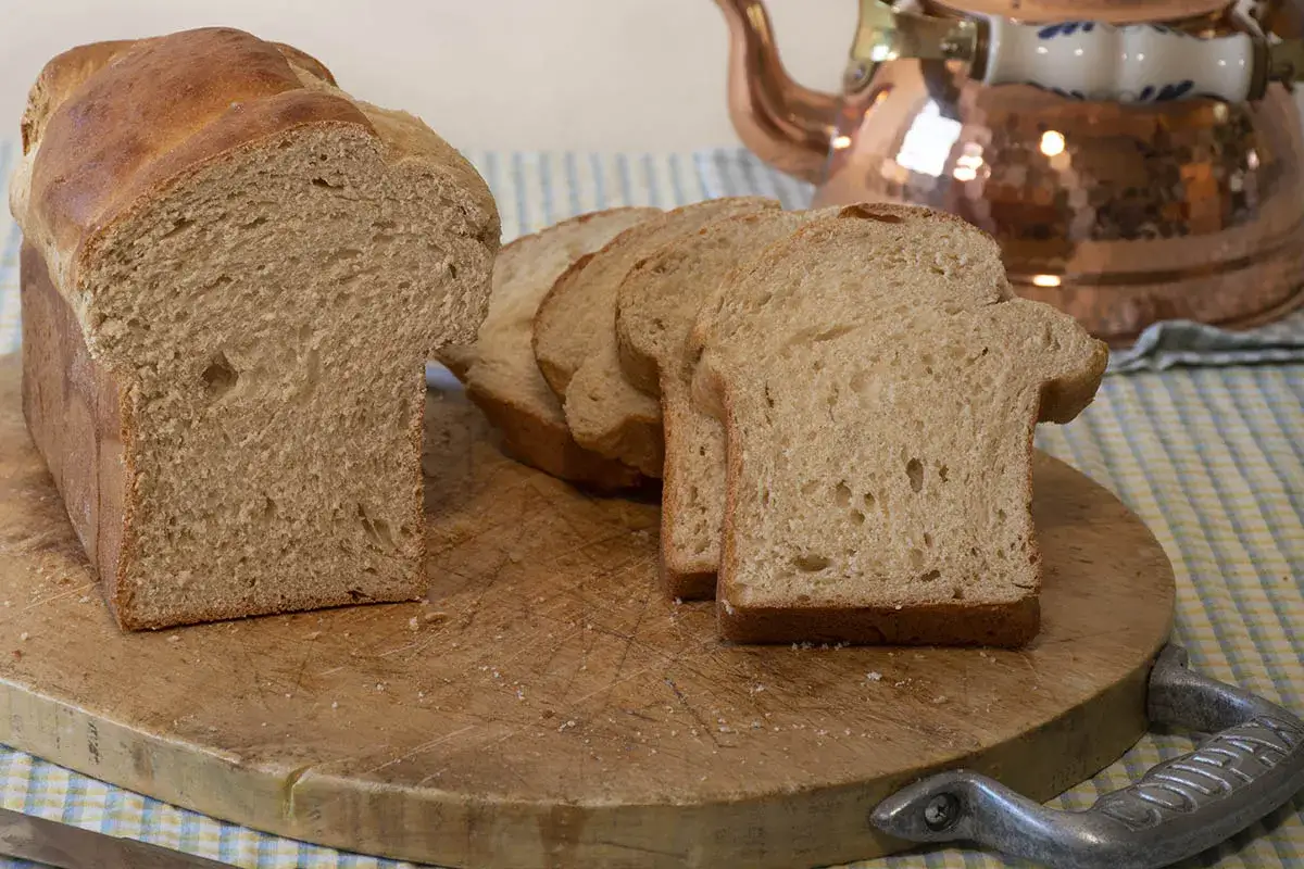 Pan para torrijas recién cortado en rebanadas sobre una tabla de madera. Una tetera de cobre brilla al fondo.