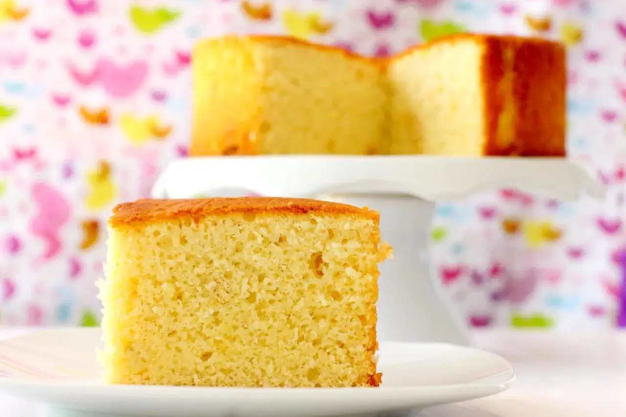Un trozo de bizcocho de yogur esponjoso y dorado sobre un plato blanco. Detrás, otro bizcocho en un soporte, con un fondo de corazones de colores.