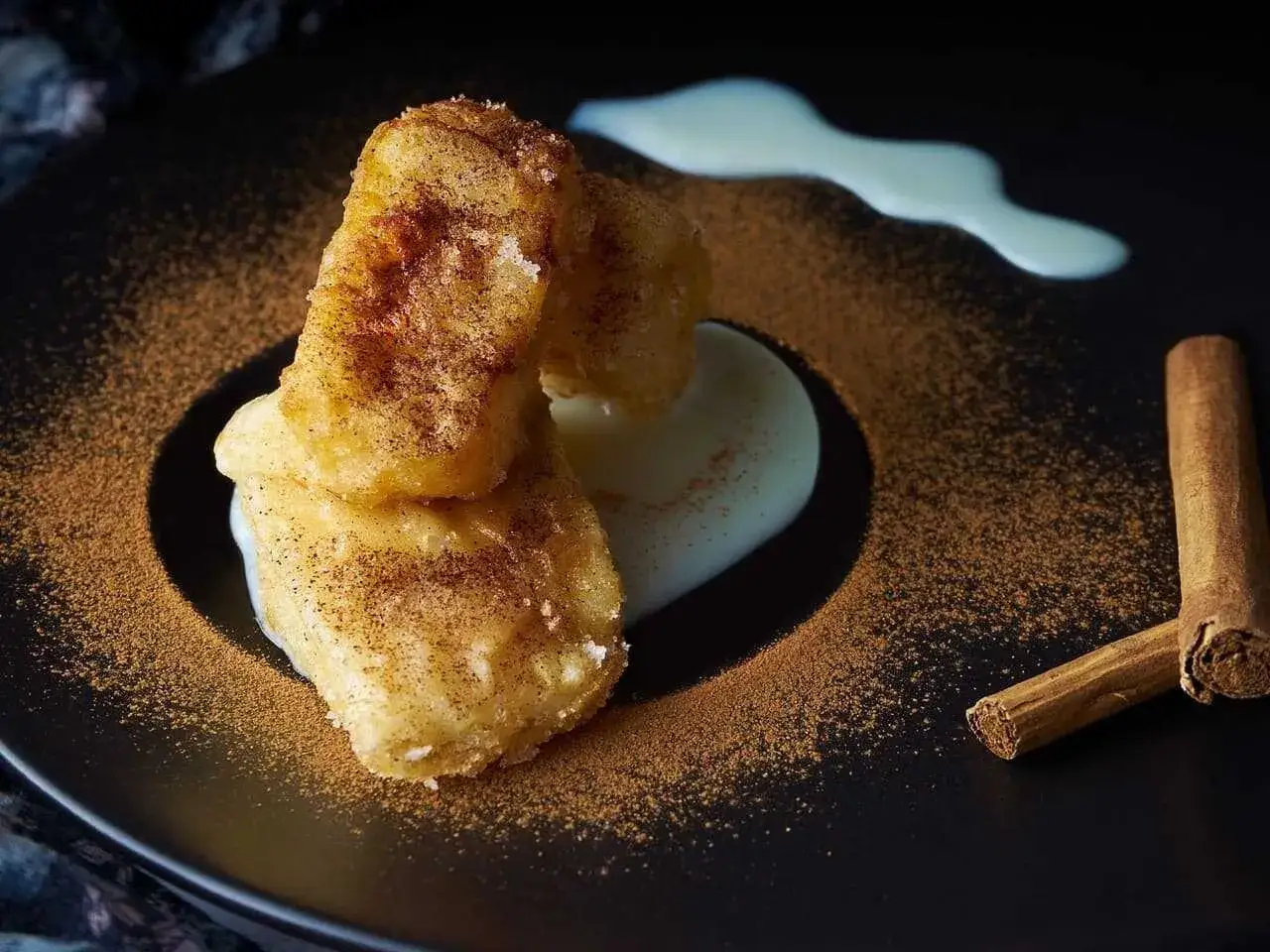 Deliciosa leche frita espolvoreada con canela, servida con crema y dos ramas de canela en una placa negra.