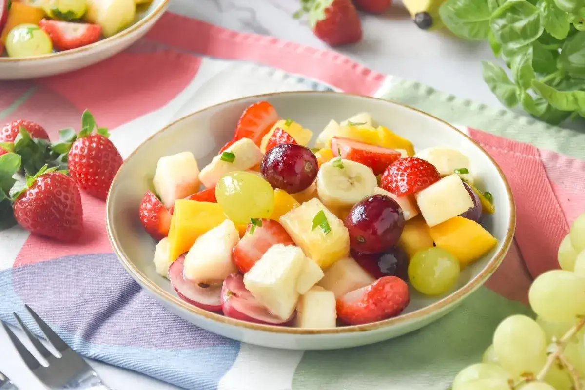 Un refrescante plato de macedonia de frutas con fresas, uvas, plátano y mango, adornado con hojas verdes.
