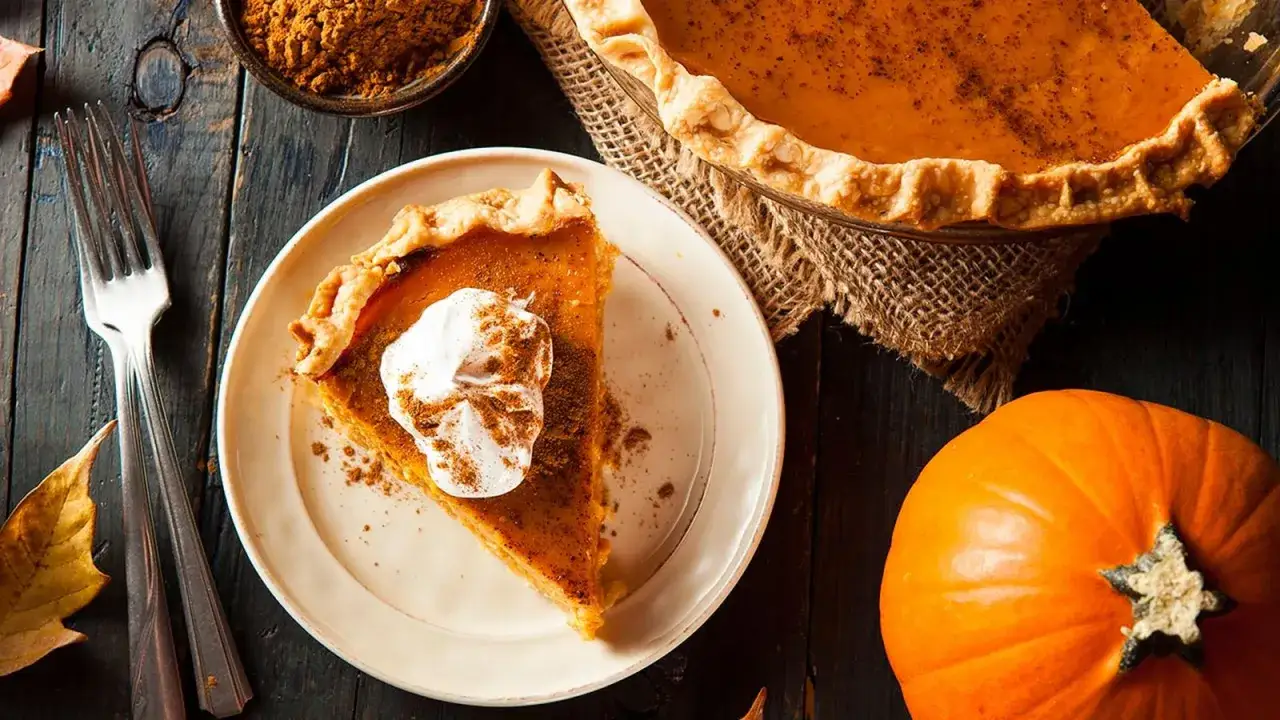 Deliciosos postres con calabaza: una rebanada de pastel de calabaza con crema batida y canela, junto a una calabaza entera y tenedores.