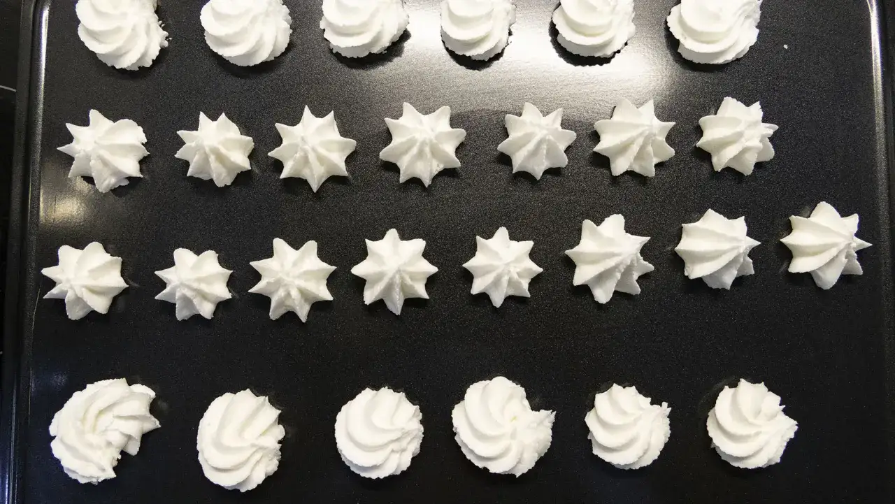 Montón de merengues con forma de estrella y espiral, listos para hornear. Parecen pequeños copos de crema chantilly.