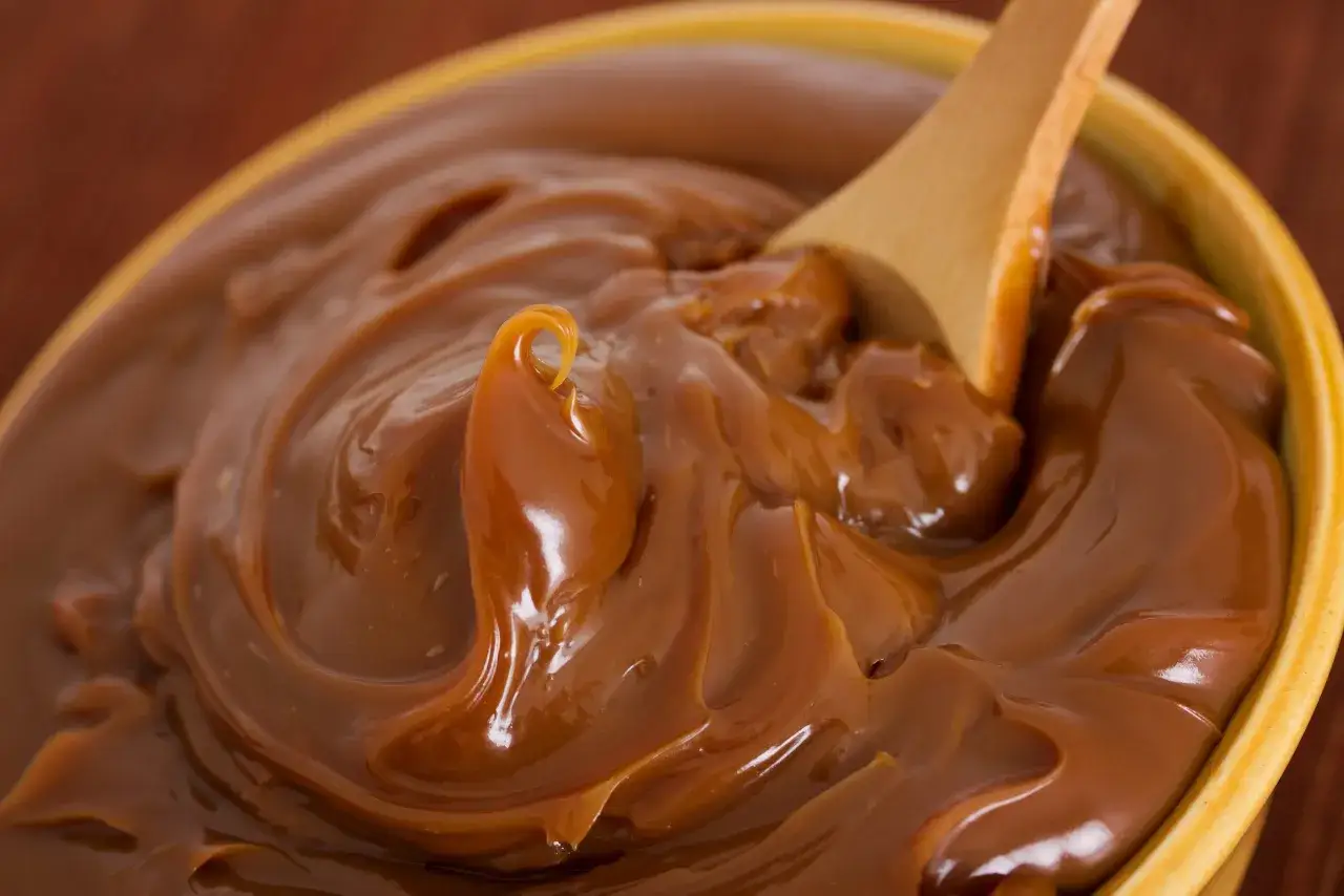 Un tazón lleno de dulce de leche cremoso, listo para ser disfrutado. Una cuchara de madera se hunde en la dulce tentación, perfecta para tu próxima dulce de leche receta.
