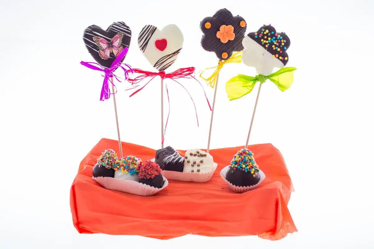 Ideas de decoracion como presentar brochetas de frutas: corazones y flores de chocolate, adornadas con cintas y chispas de colores, sobre una base naranja.