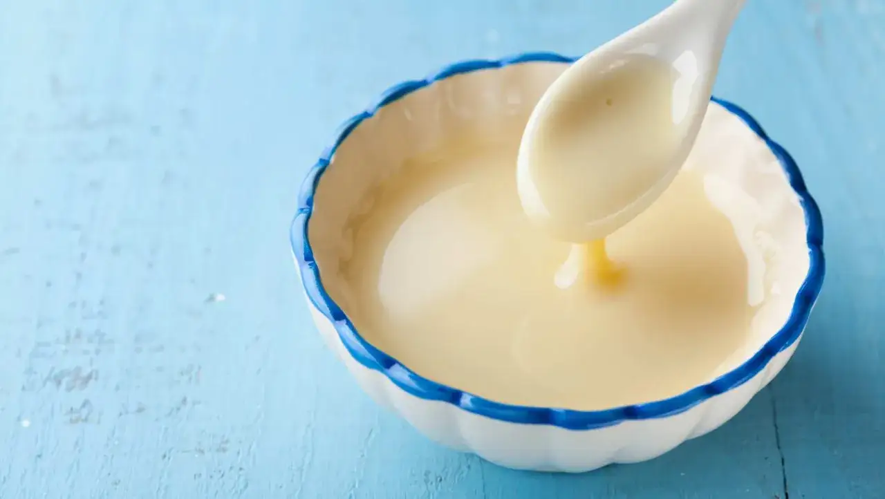 Cuchara con leche evaporada, cremosa y espesa, cayendo en un cuenco blanco con borde azul.