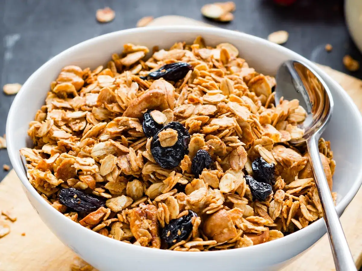 Tazón blanco lleno de granola casera crujiente con pasas y nueces. Una cuchara plateada descansa en el borde.