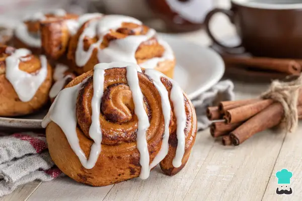 Rollo de canela cubierto de glaseado blanco, con canela en rama atada y una taza de café al fondo.