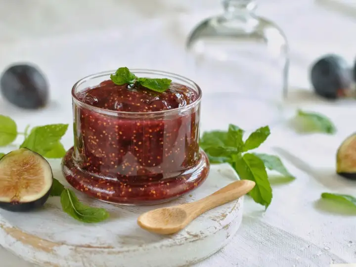 Tarro de mermelada de higos casera, decorado con hojas de menta. Higos frescos y una cuchara de madera completan la escena.
